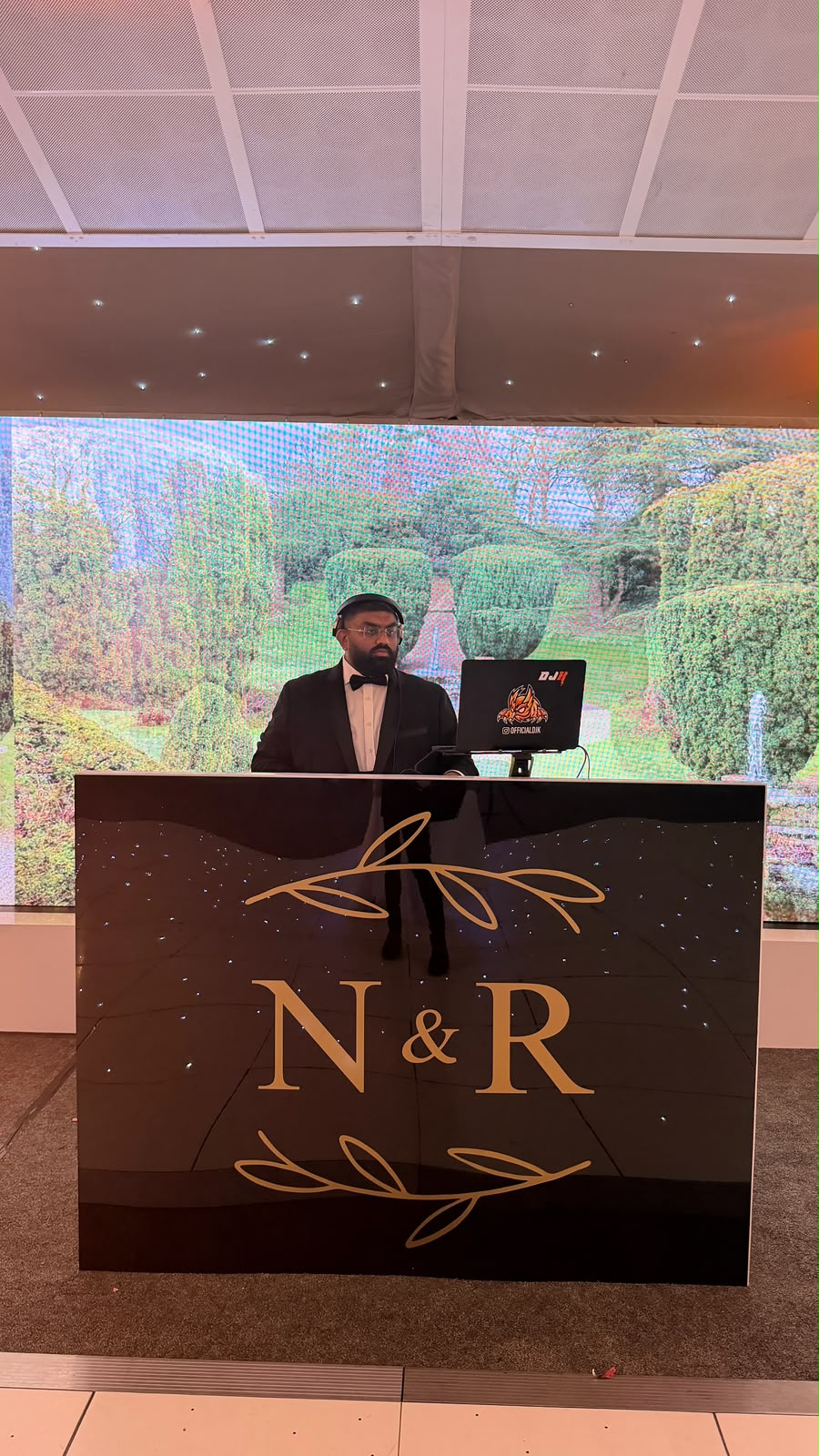 DJK behind the decks at a high-end event with live screens behind him.